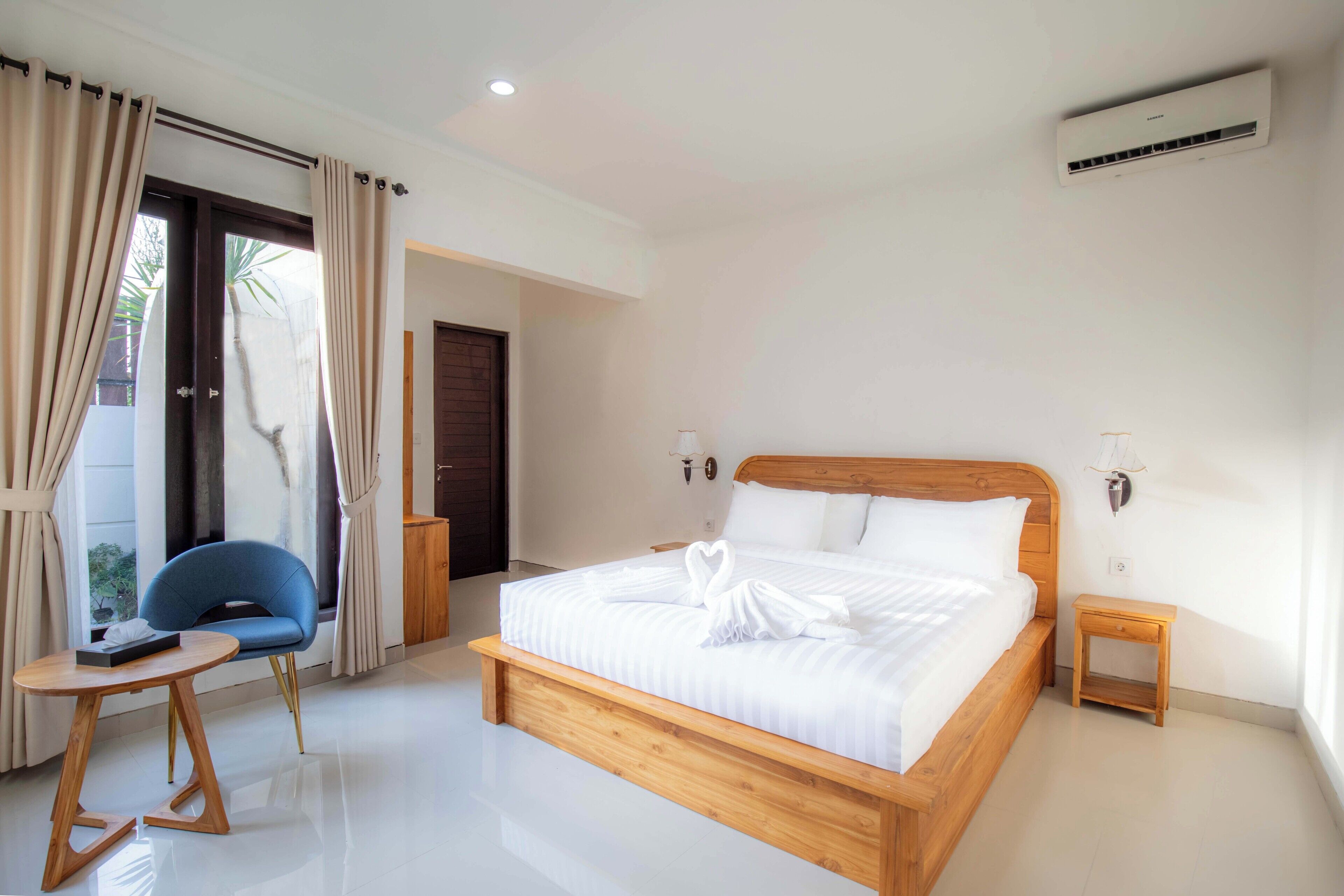 A modern hotel room with a wooden bed, a small round table, a blue chair, and a view through a glass door.