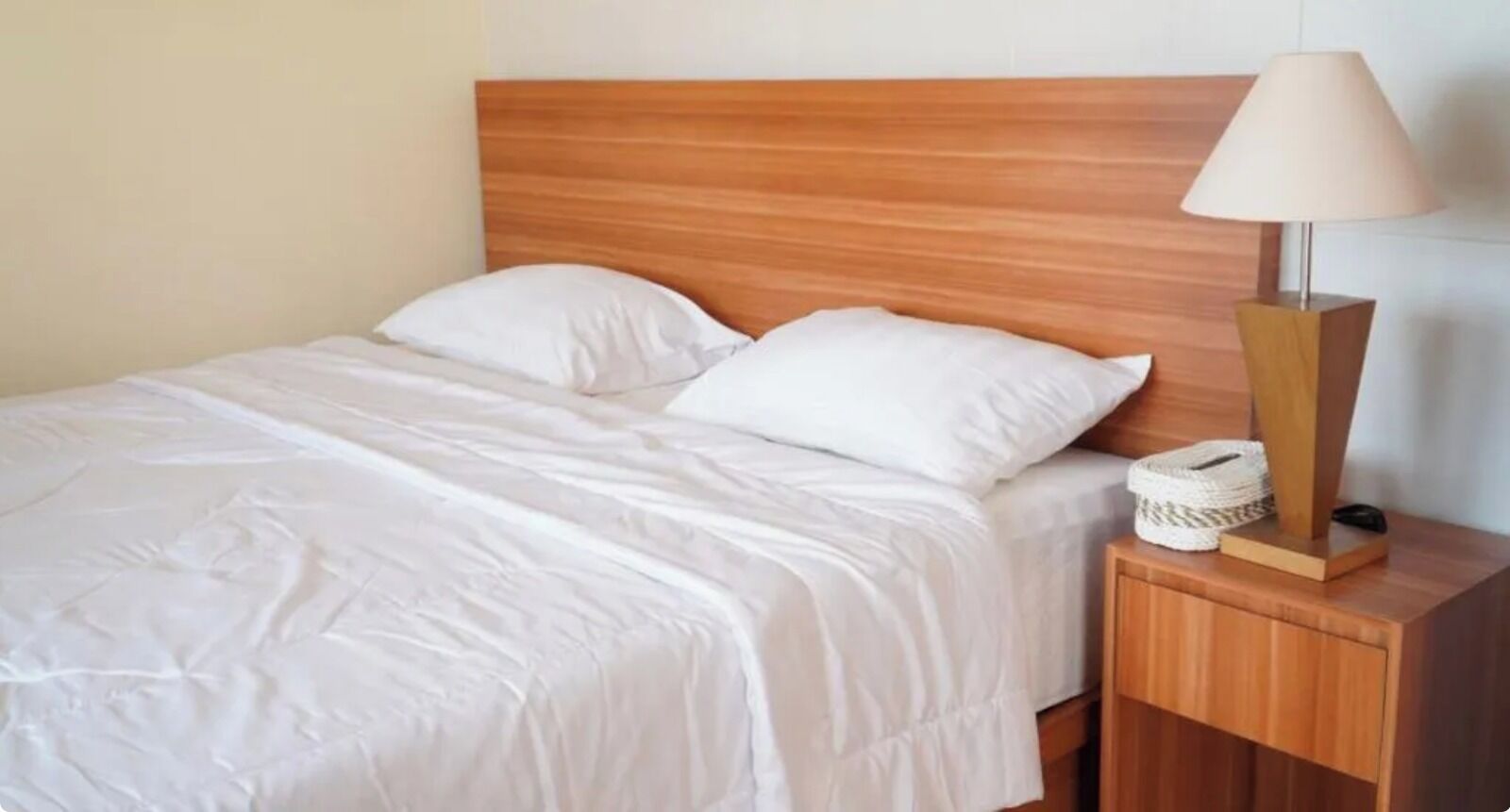 A neatly made bed with white linens, a wooden headboard, and a bedside table with a lamp and a woven basket.