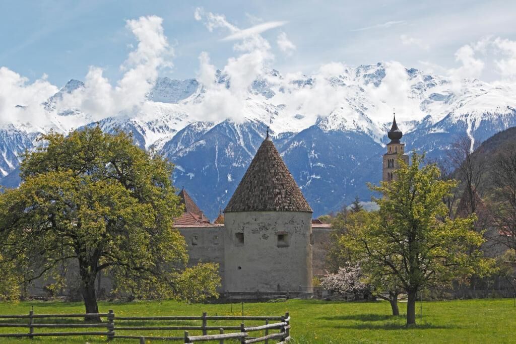 A historic building with a conical roof, surrounded by a wooden fence and trees, with snow-capped mountains in the background.