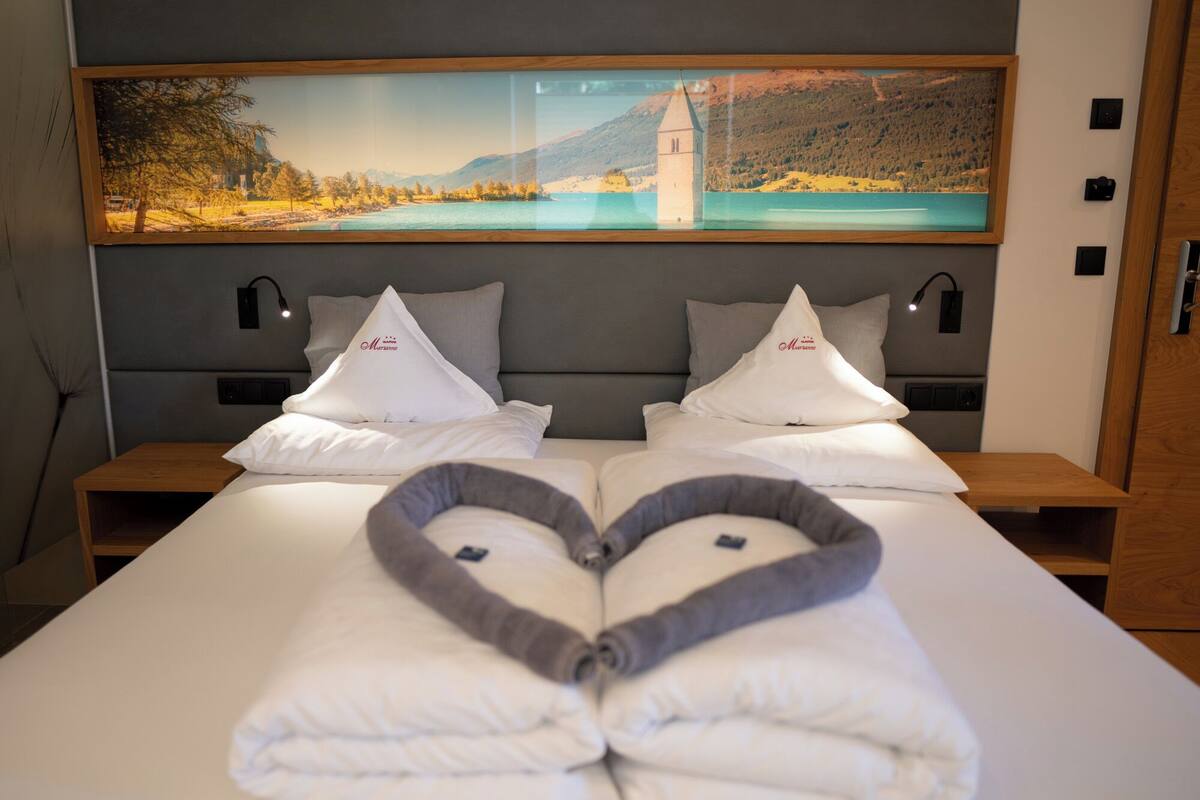 Habitación de hotel con una cama, dos mesitas de noche, un cuadro grande de un paisaje y una disposición de toallas con forma de corazón.