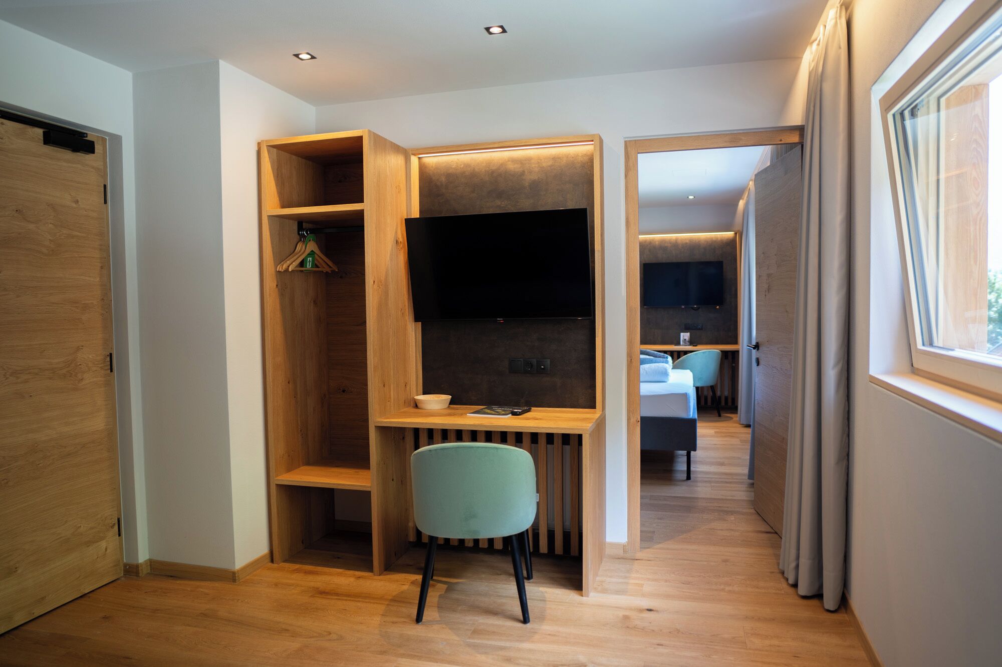 A modern, open-plan room with a wooden desk, a chair, a television, and a view of another room with a bed and a table.