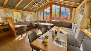 Un comedor moderno con mesas y sillas de madera, ventanas amplias con vistas a árboles y montañas, y un techo alto con vigas a la vista.