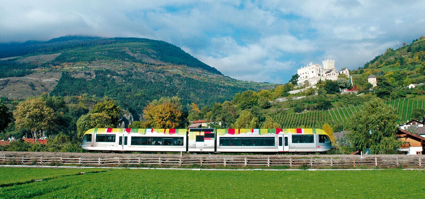 Ein Zug mit bunten Wimpeln durch eine ländliche Landschaft, im Hintergrund ein Schloss.