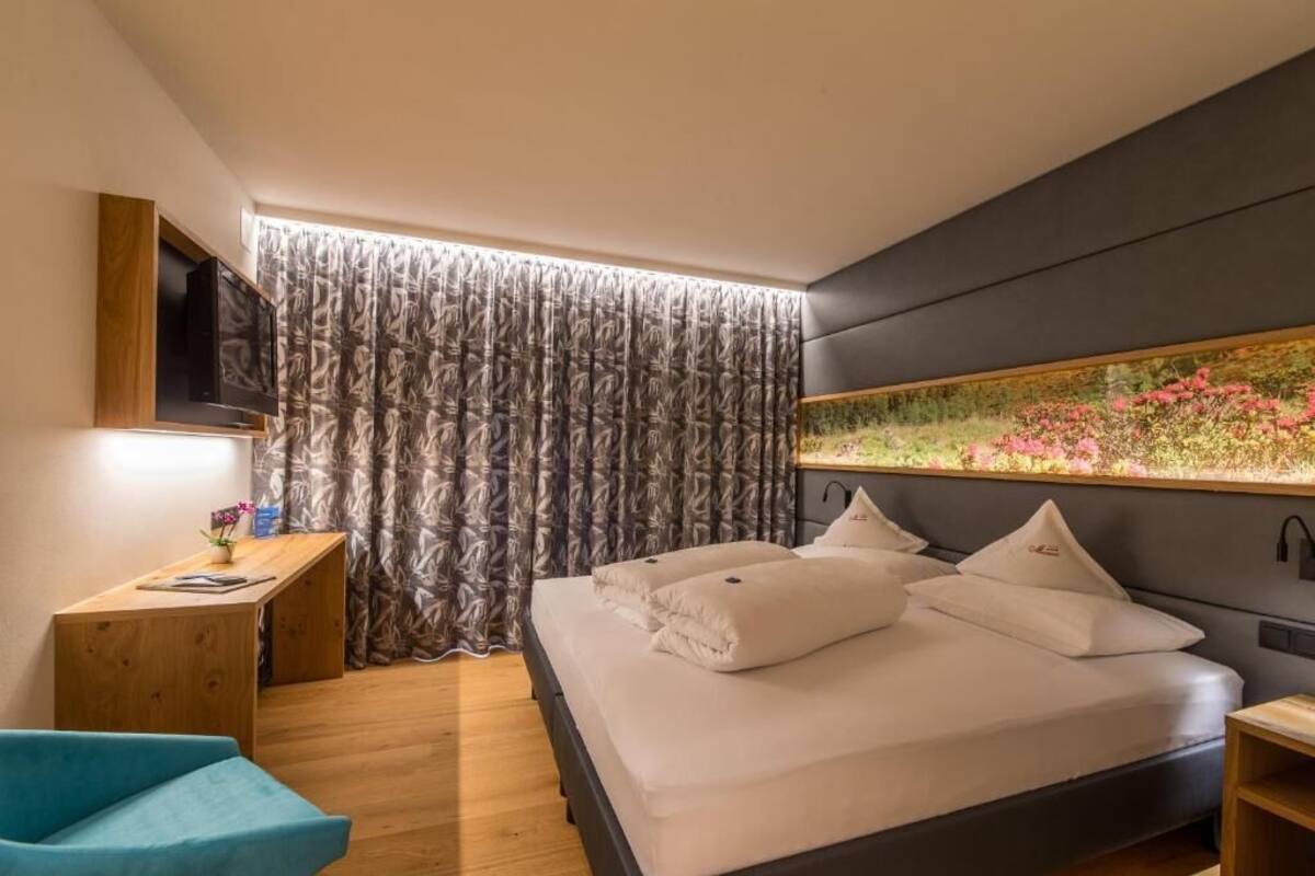Una habitación de hotel moderna con una cama, un televisor montado en la pared, un escritorio y un ventanal con cortinas.