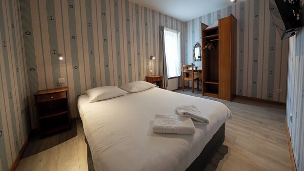 A neatly made bed with pillows and a folded towel, a wooden nightstand, a chair, and a wooden wardrobe in a room with striped wallpaper. - Hôtel Le Relais Du Canalou (Digoin)