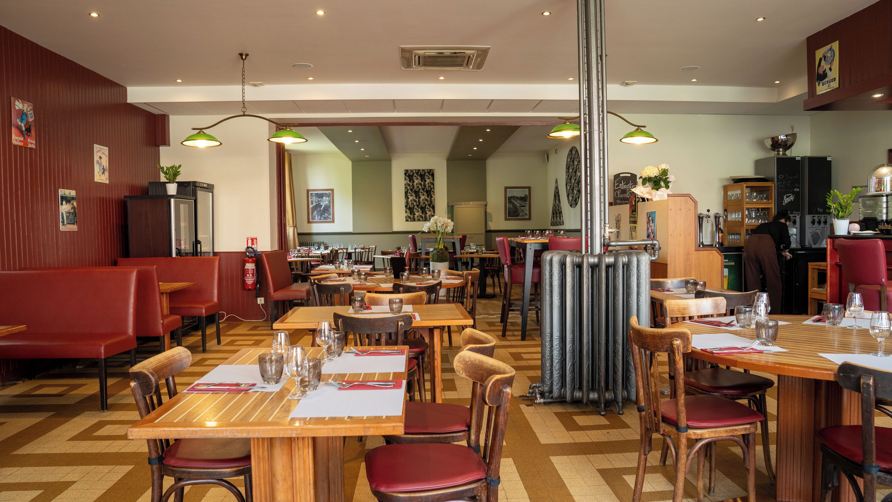 A restaurant with wooden tables and red upholstered booths, featuring a central decorative radiator, multiple hanging green lamps, and a visible fire extinguisher.