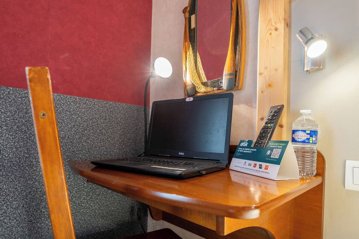 Un bureau en bois avec un ordinateur portable, un miroir, une télécommande et une bouteille d’eau.