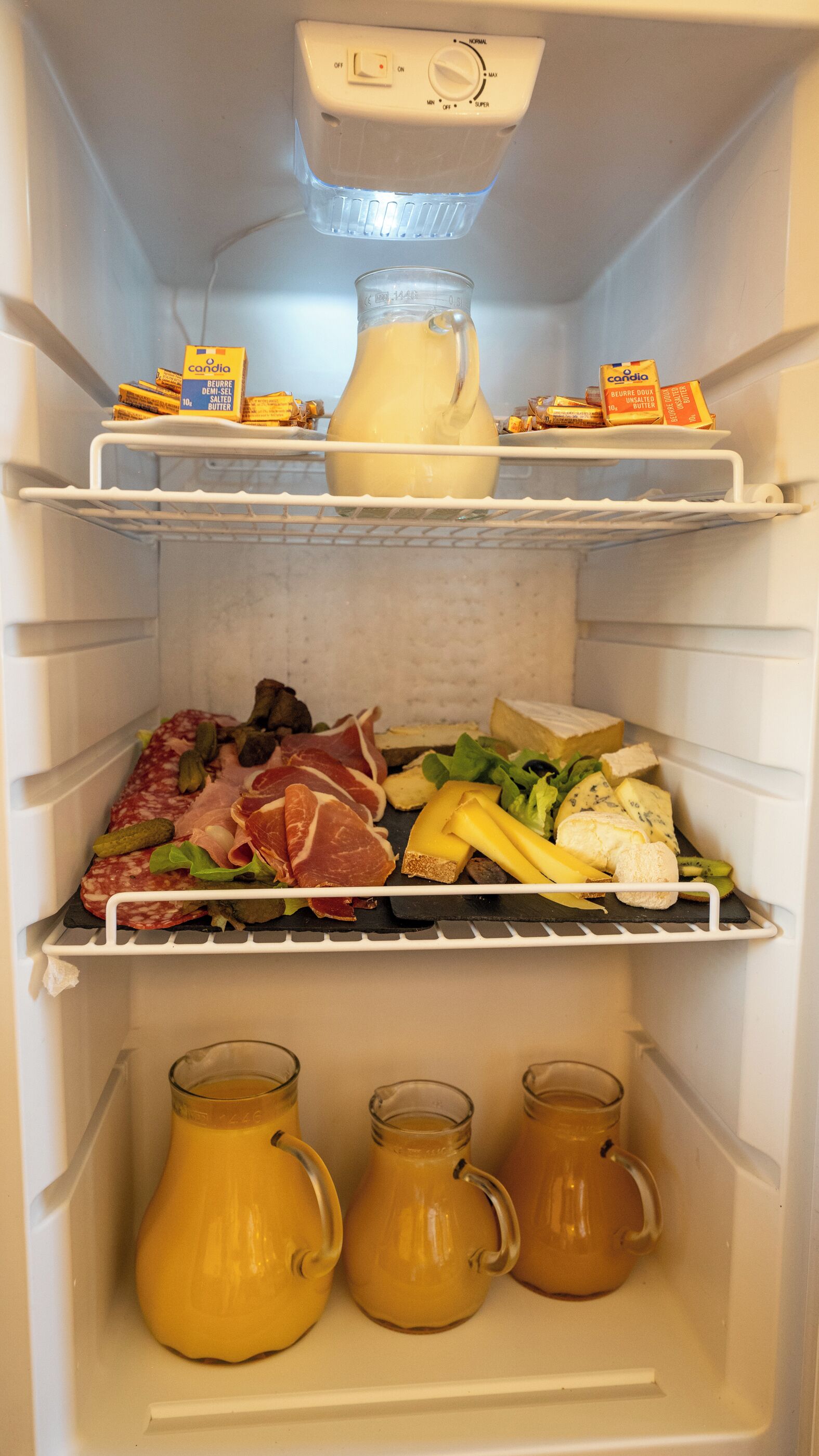 Un refrigerador abierto con repisas llenas de diversos alimentos, como queso, carnes y condimentos.