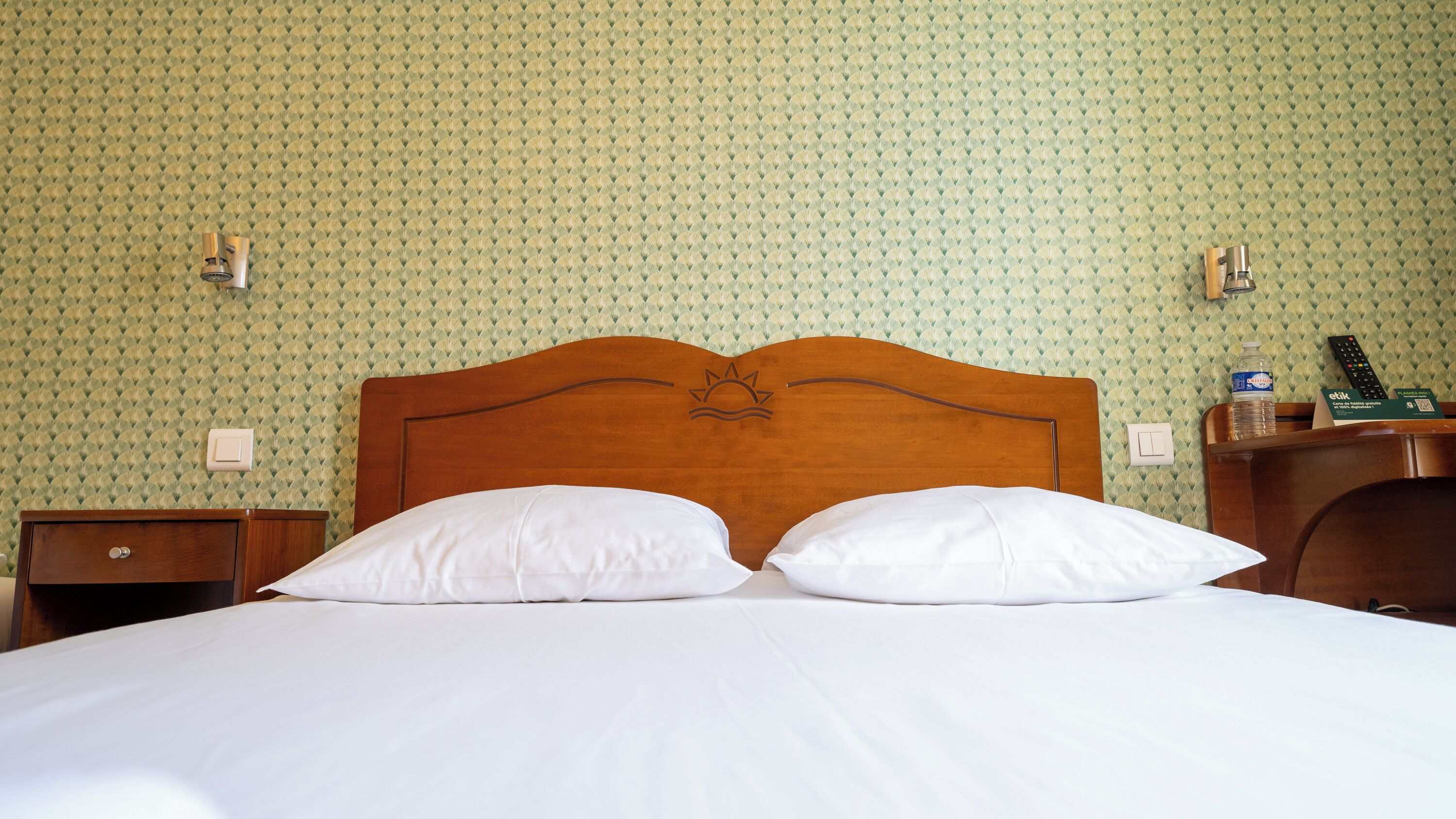 Ein Hotelzimmer mit einem hölzernen Kopfteil, zwei weißen Kissen, einem Nachttisch aus Holz, einer Nachttischlampe und einem an der Wand montierten Lichtschalter.