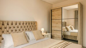 Quarto com cabeceira estofada, cama com roupa de cama bege, mesa de cabeceira com abajur e guarda-roupa espelhado.