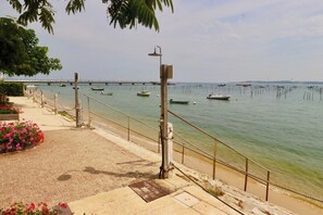 A coastal walkway with a railing, a pier in the distance, and boats on the water.