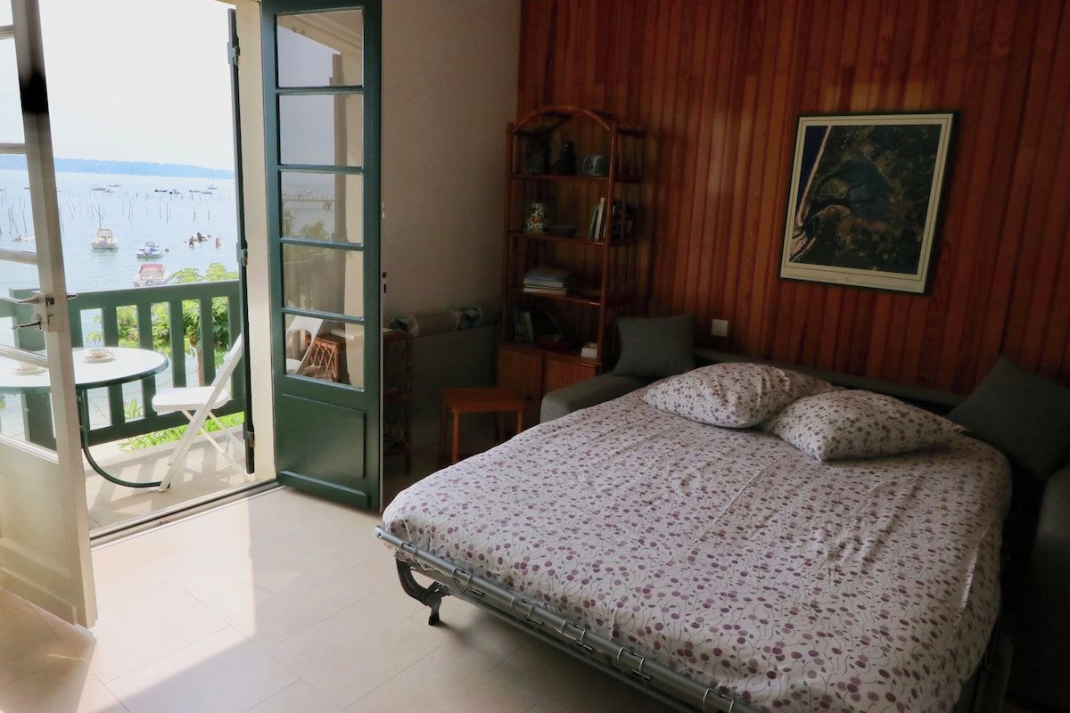 A bedroom with a bed, a wooden bookshelf, a chair, and a view of the sea through an open door.