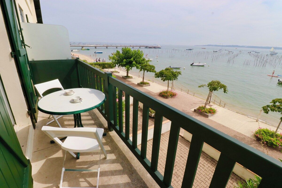 A balcony with a table and chairs, overlooking a calm sea with boats and a pier in the distance.