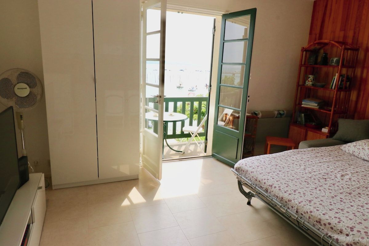 A bedroom with a bed, a fan, a wooden bookshelf, a small table, and a balcony with a view of boats.