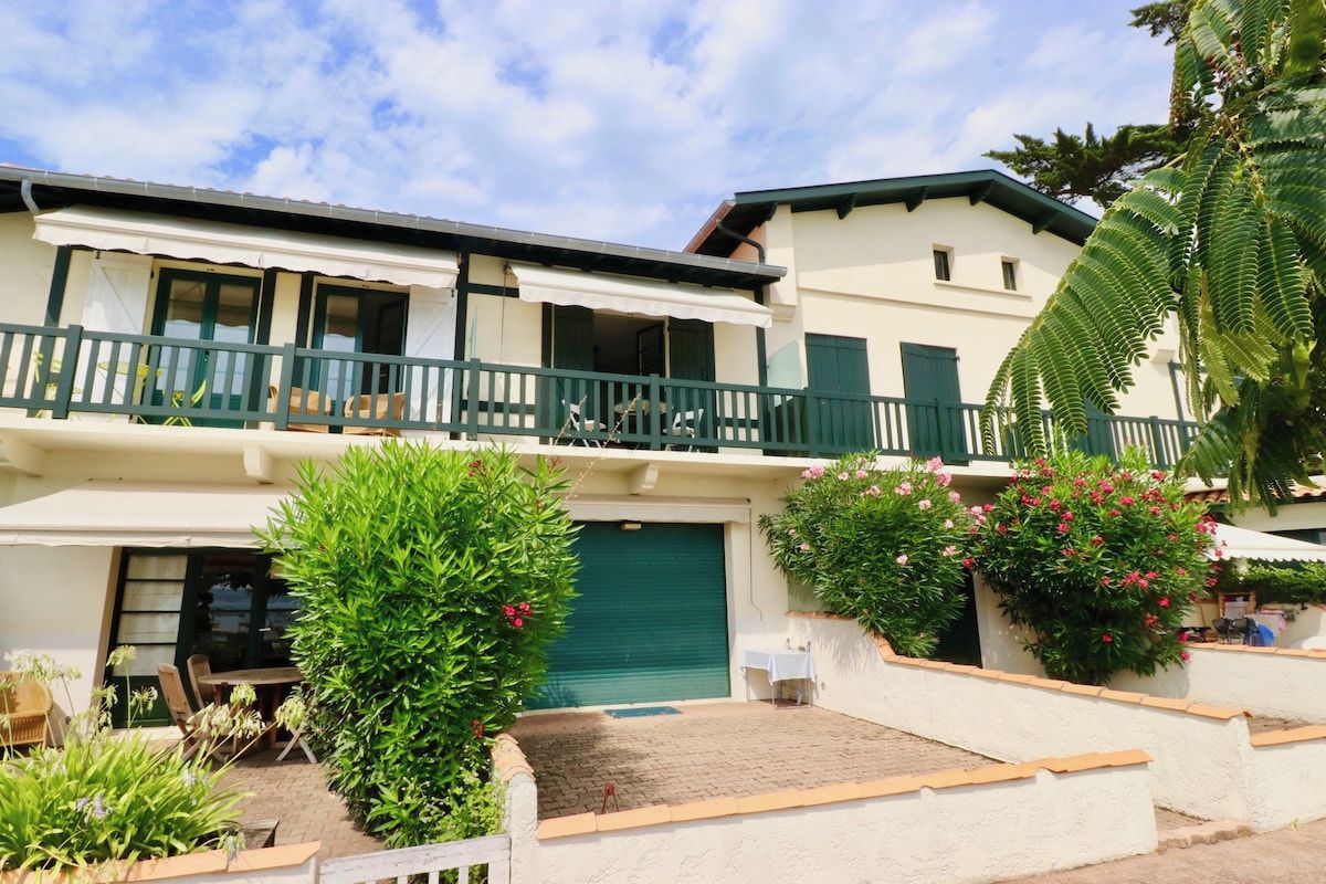 A two-story building with a balcony, a garage, and a garden area with plants and flowers.
