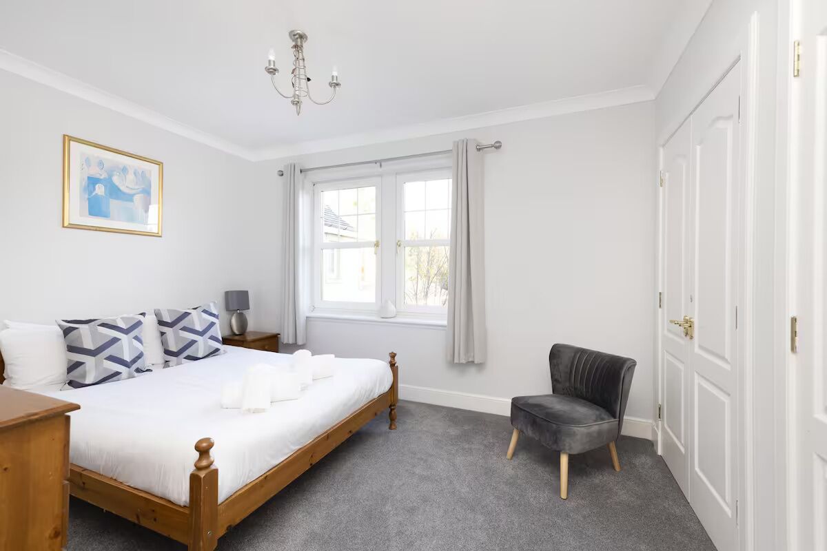A bedroom with a wooden bed, a chair, a nightstand, a lamp, a window with curtains, and a chandelier.