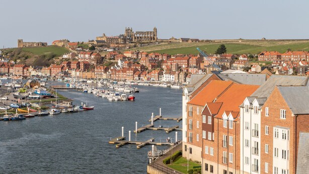 Un pueblo costero con edificios históricos, un puerto deportivo y un castillo al fondo.