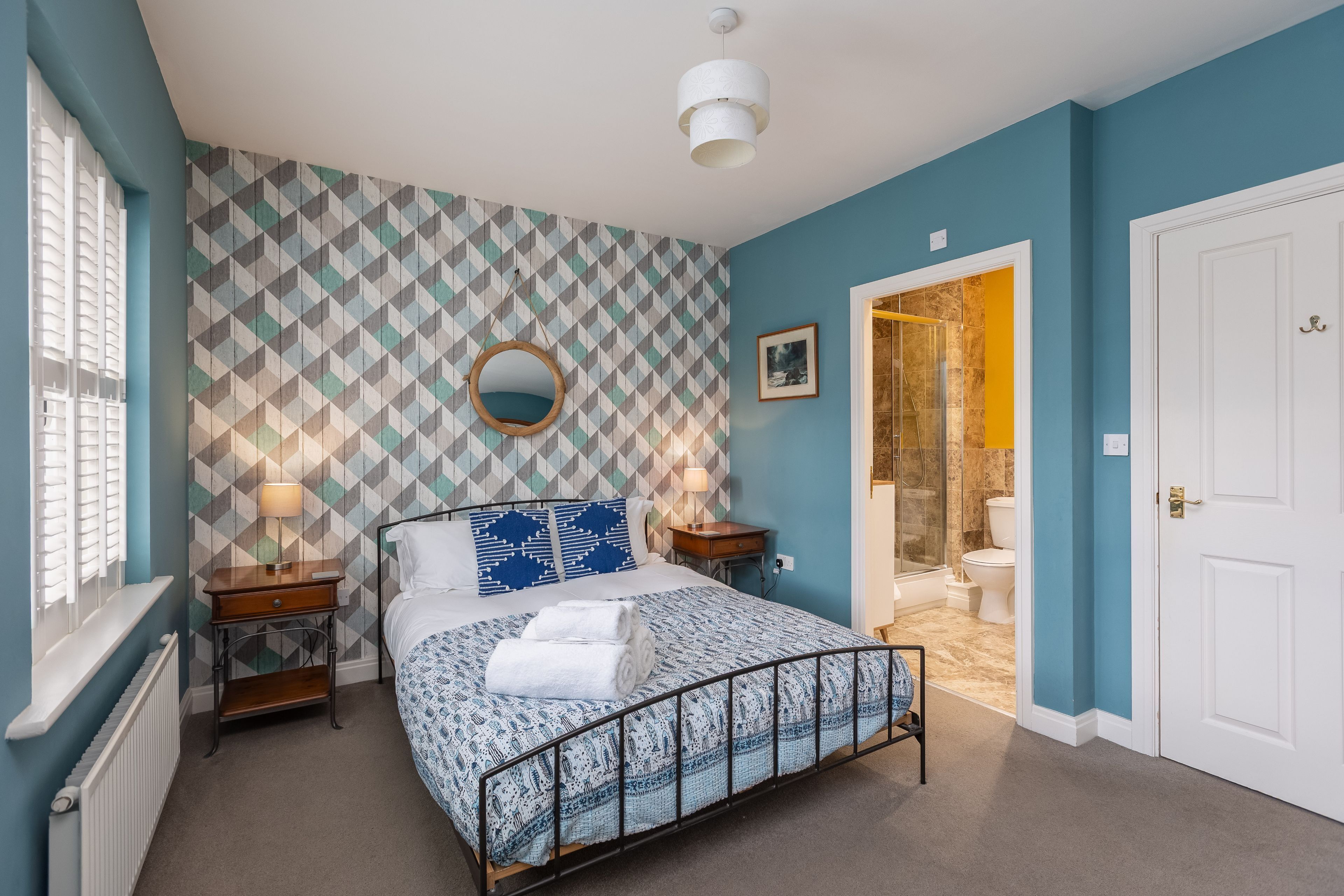 Un dormitorio con pared decorada, una cama con estructura metálica, mesitas de noche con lámparas y vista al baño.