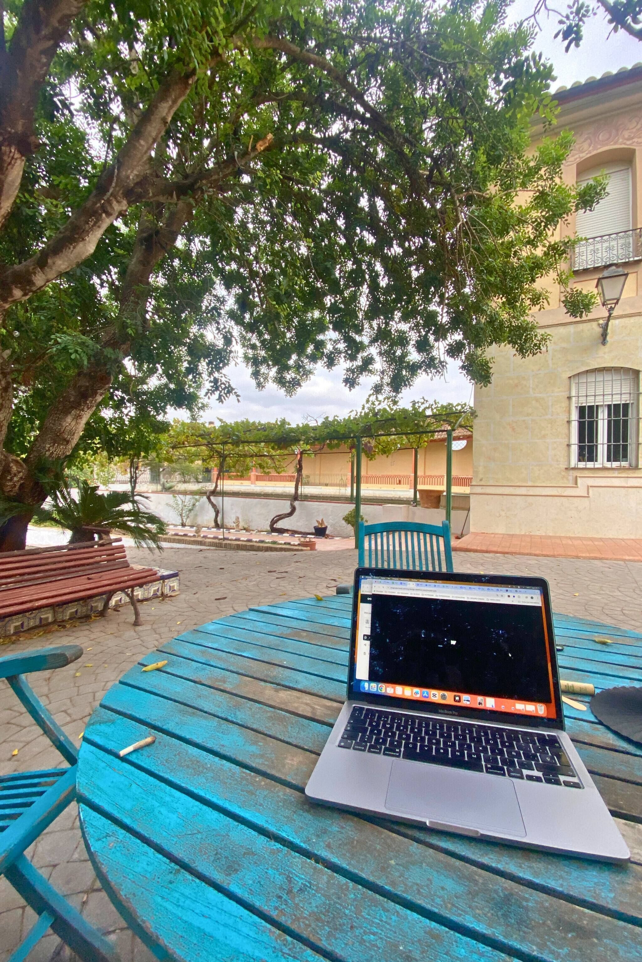 Una computadora portátil sobre una mesa azul en un entorno exterior, con un árbol y un edificio al fondo.