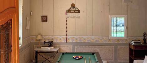 A room with a pool table, wooden beams on the ceiling, a chandelier, and a window with a view.