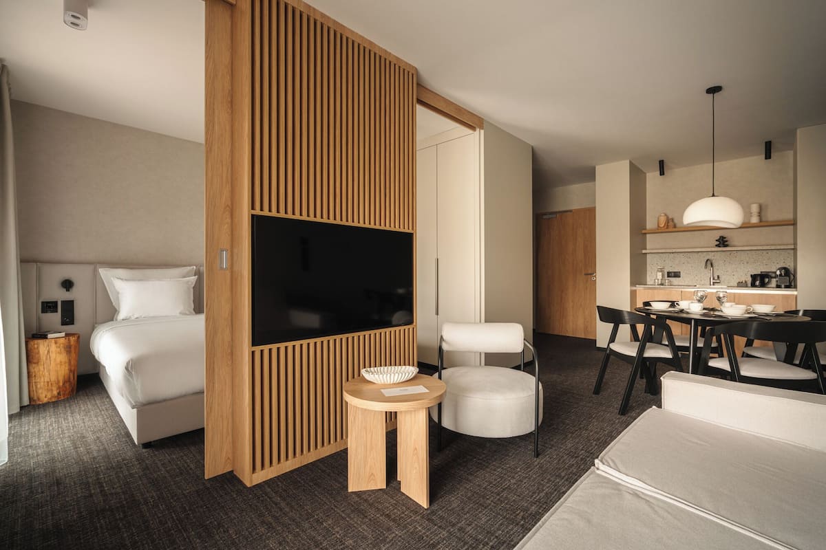 Quarto de hotel moderno com cama, TV, divisória de madeira, mesa redonda e cozinha compacta.