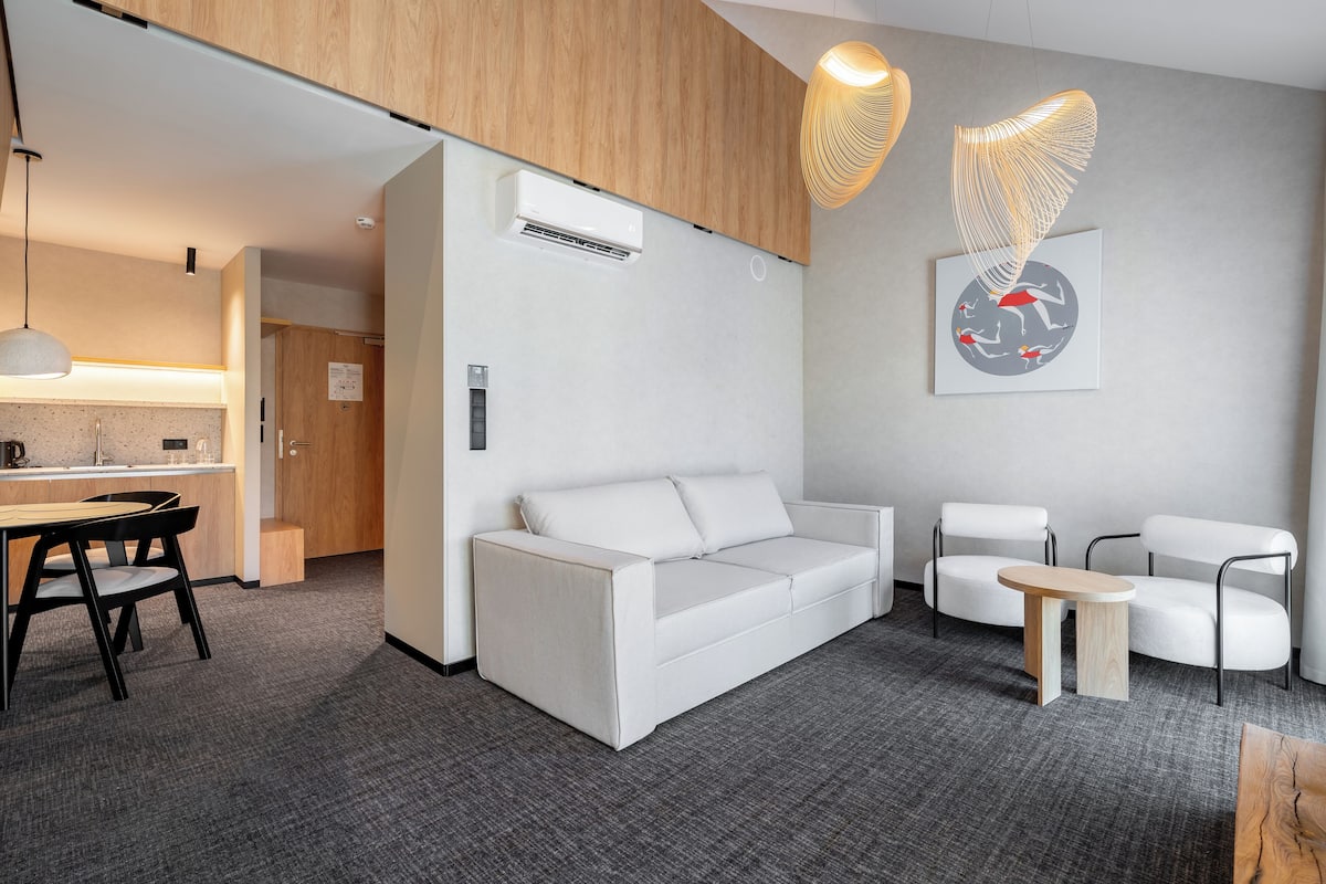 Quarto de hotel moderno com sofá branco, pequena mesa lateral de madeira e área de refeições com mesa e cadeiras.