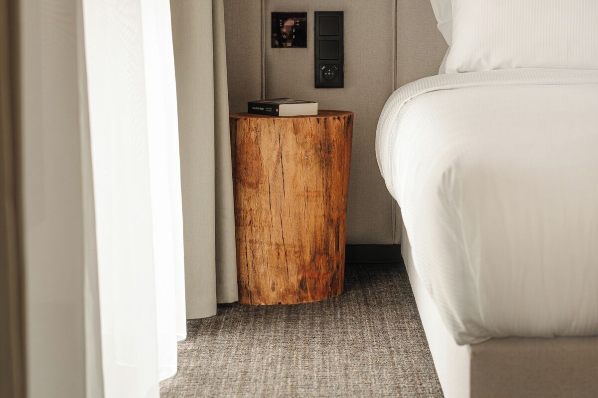 Una habitación de hotel con una cama, una mesita de madera y un panel de control en la pared, además de una cortina.