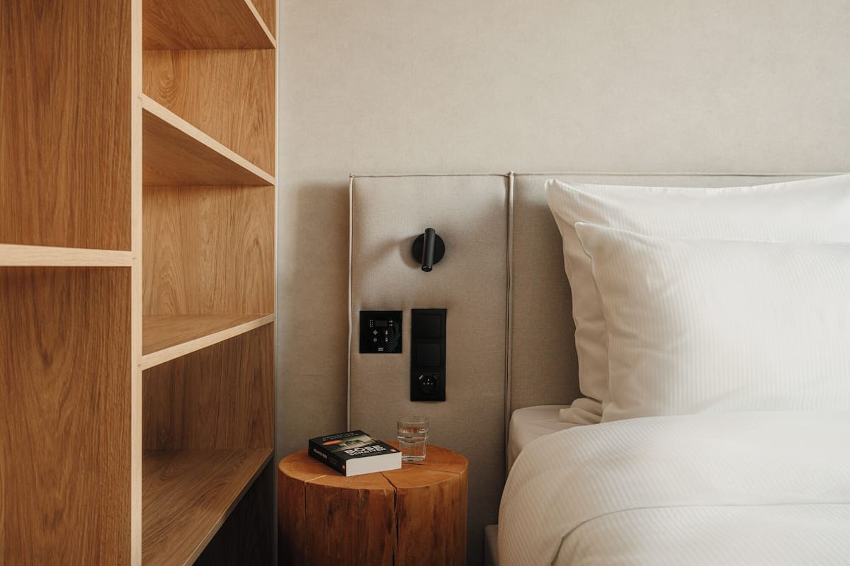 Quarto de hotel com guarda-roupa de madeira, mesa de cabeceira com um livro e um copo, e cama com roupa de cama branca.
