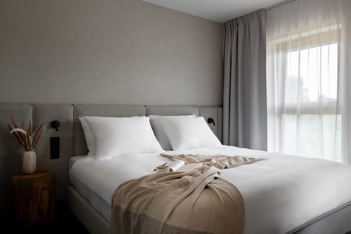 Quarto moderno com cama grande, mesa de cabeceira com um vaso e vista para o exterior através de cortinas leves.