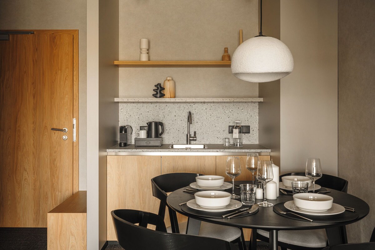 A compact kitchen with a dining area. It includes a sink, a coffee maker, a kettle, and a hanging light fixture.