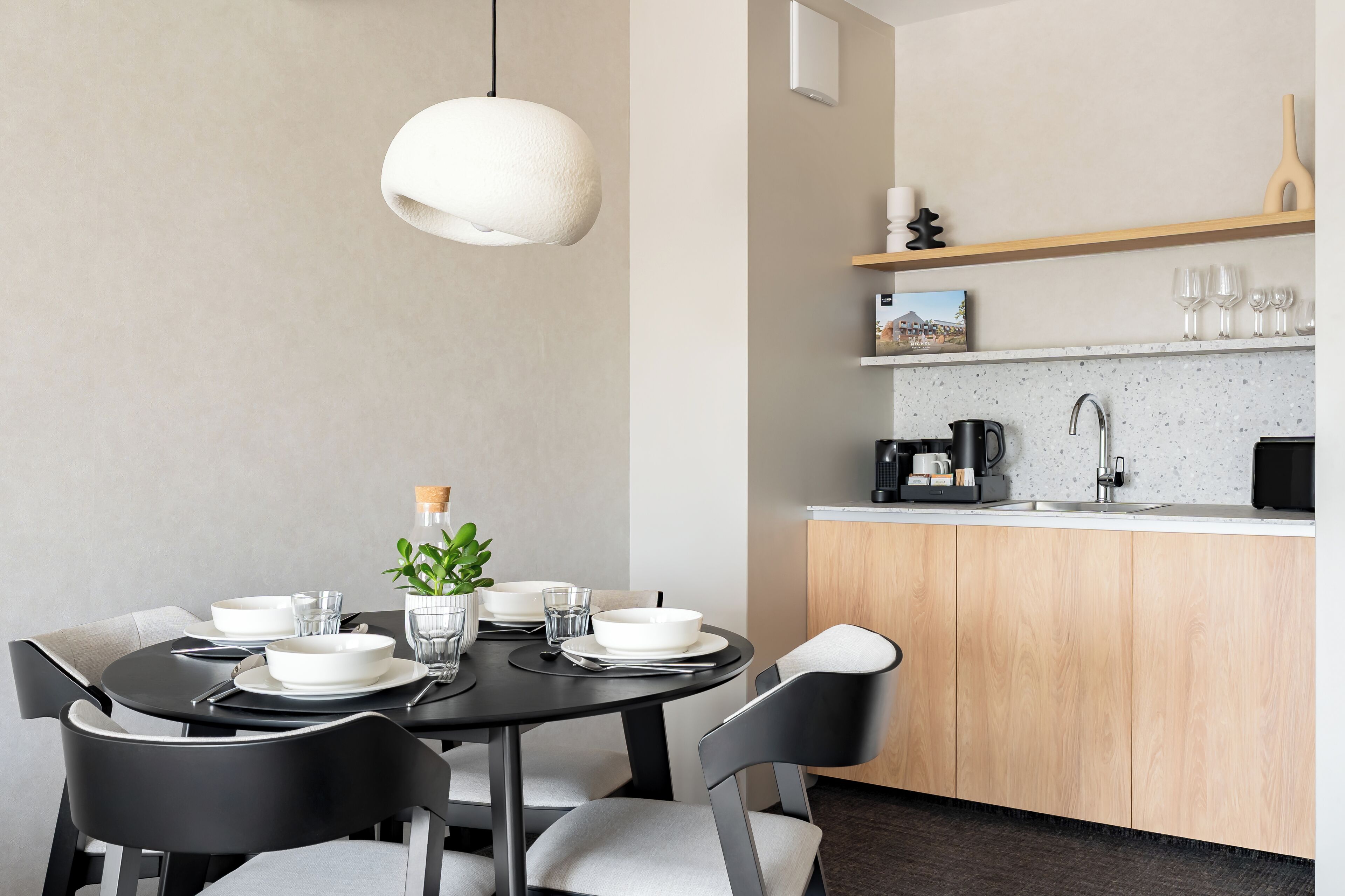 Un espace repas moderne avec une table ronde pour quatre personnes, un luminaire suspendu et un coin cuisine équipé d’un four à micro-ondes, d’une machine à café et d’une étagère en bois au-dessus de l’évier.