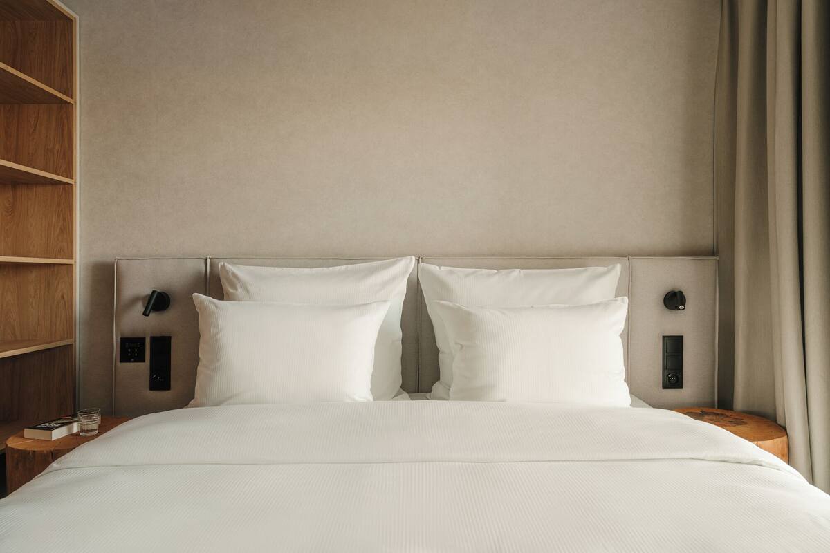 Quarto de hotel com cama, duas mesas de cabeceira, um criado-mudo, prateleira de madeira e painel de controle fixado na parede.