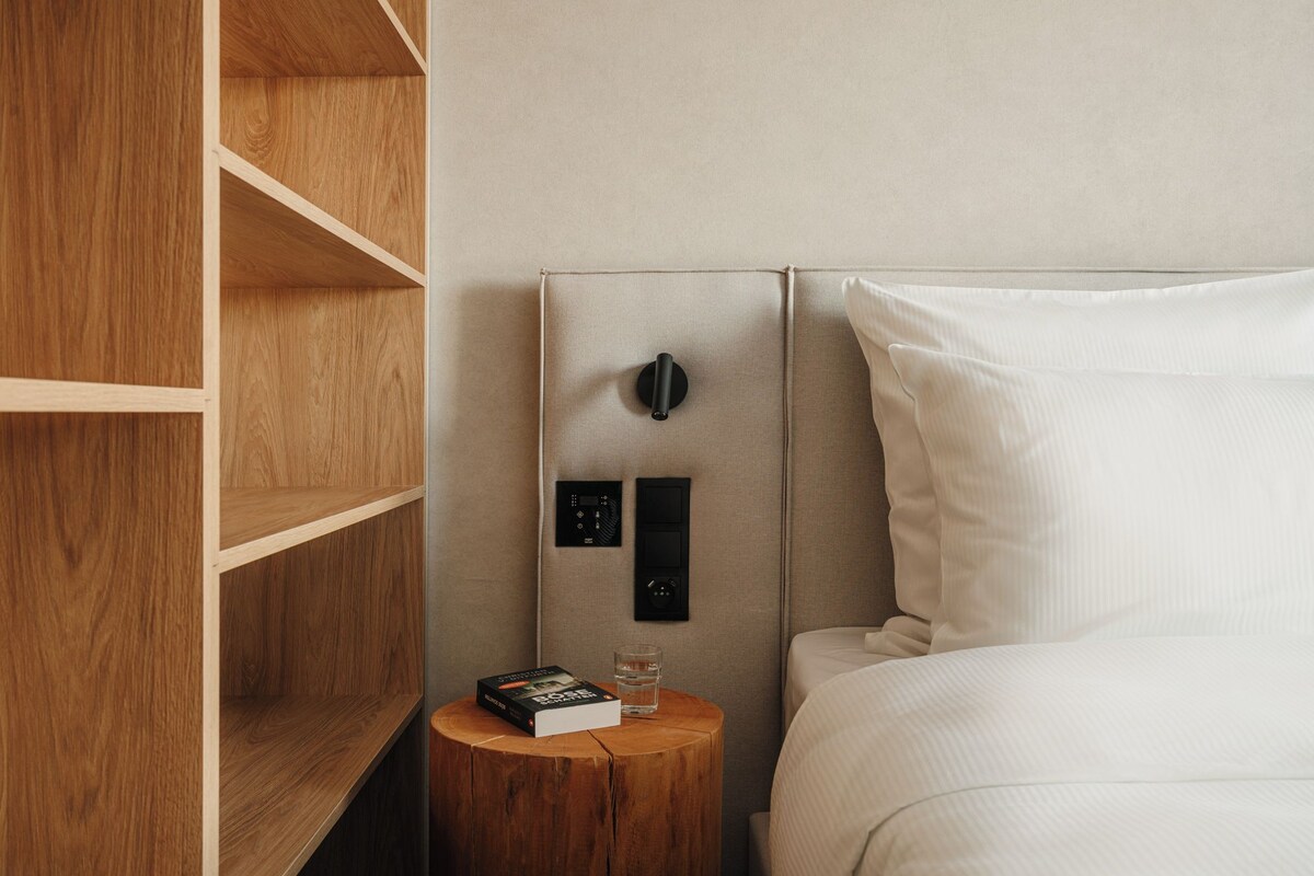 A hotel room with a wooden wardrobe, a bedside table with a book and glass, and a bed with white bedding.