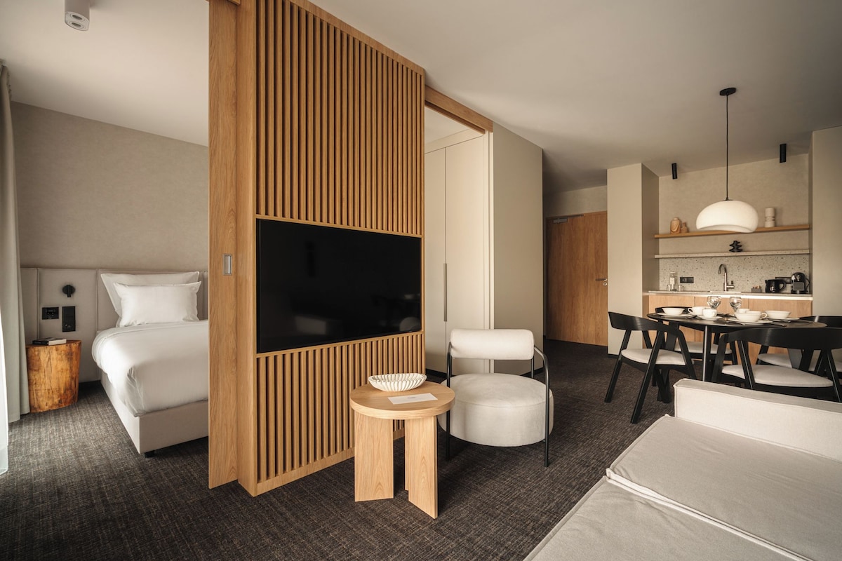 A modern hotel room with a bed, TV, wooden partition, round table, and a kitchenette.