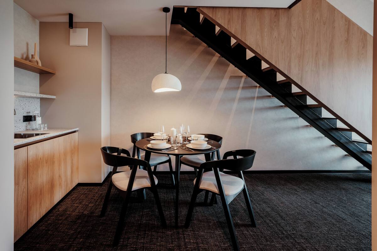 Uma sala de jantar moderna com uma mesa redonda posta para seis pessoas, uma escada e uma cozinha com armários de madeira e bancada.