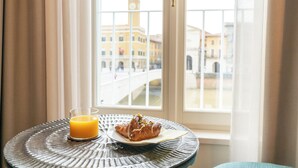 A breakfast table with a croissant and a glass of orange juice by a window with a view of a bridge and buildings. - Anna Teresa Luxury (Pisa)