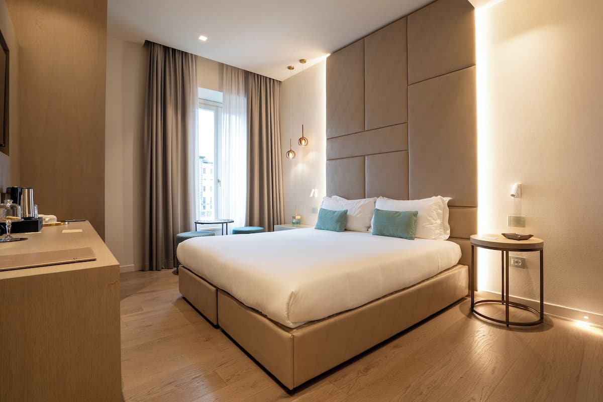 A modern hotel room with a large bed, a bedside table, a desk, and a window with curtains.