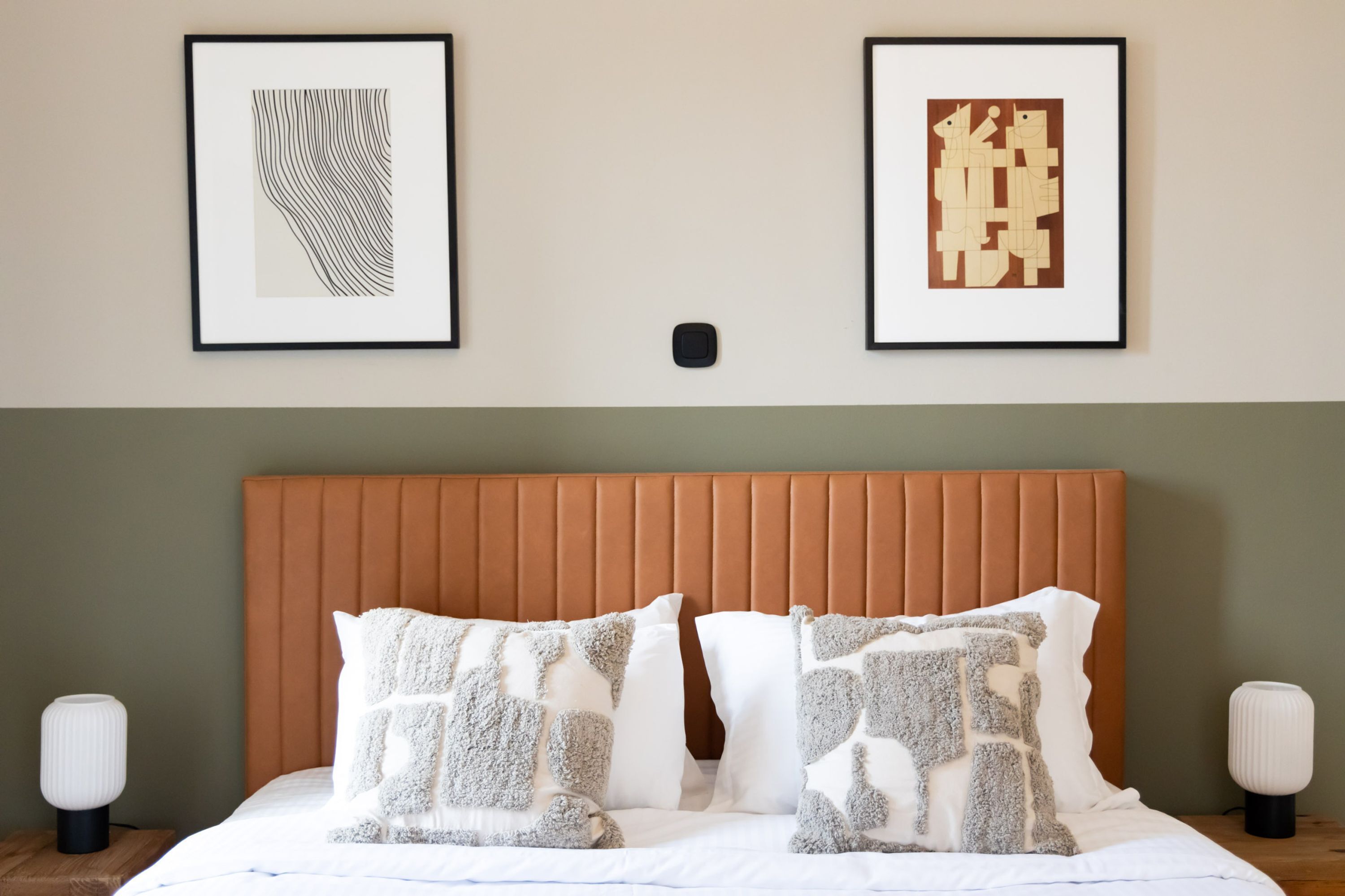 A neatly made bed with patterned pillows, a padded headboard, and two framed artworks on the wall.