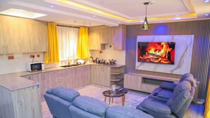 Una sala moderna con un televisor grande, un sofá, una mesa de centro y una cocina con armarios de madera y cortinas amarillas.