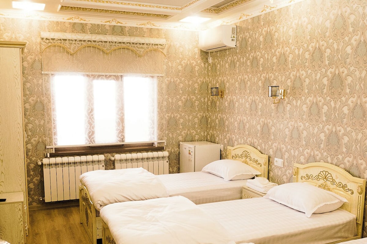 A hotel room with three beds, a radiator, a small refrigerator, and ornate wallpaper.