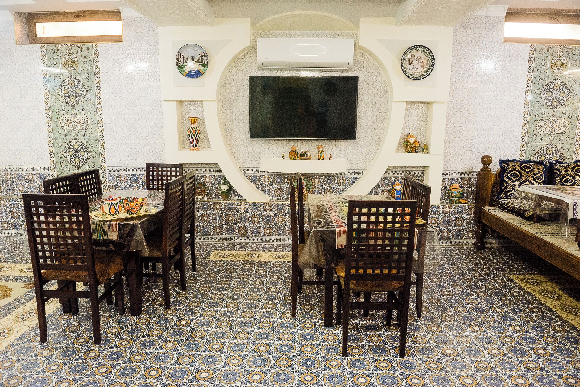 A dining area with a TV mounted on the wall, surrounded by patterned tiles and wooden chairs.