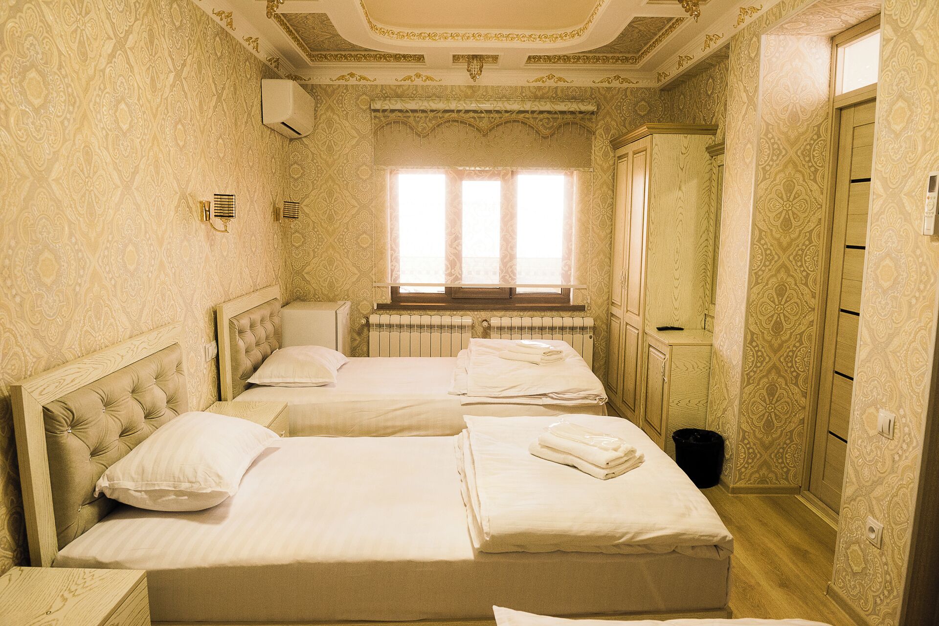 A hotel room with two beds, a radiator, a window with curtains, and patterned wallpaper.
