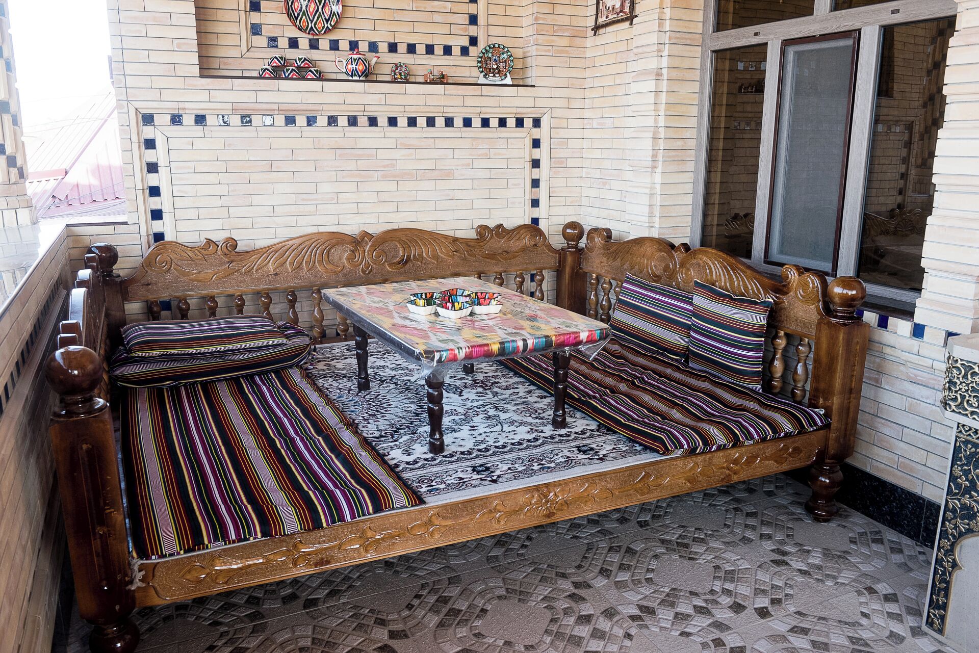 A wooden bench with a table in the center, patterned cushions, and a decorative wall with tiles and a window.