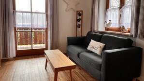 A cozy living room with a wooden floor, a grey sofa with a pillow, a wooden coffee table, and a large window with curtains. - Fewo Sonnenglück - dreamlike 2-room apartment for 1-2 persons with corner balcony (Schönau am Königssee)
