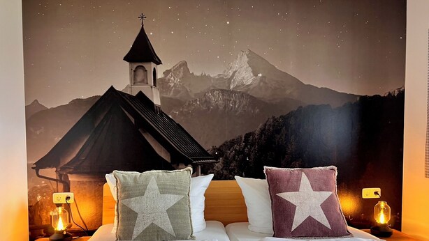 A bed with white bedding and star-patterned pillows, set against a mural of a mountain church and mountains. - Fewo Sonnenglück - dreamlike 2-room apartment for 1-2 persons with corner balcony (Schönau am Königssee)