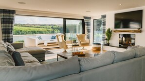Un salon moderne avec un grand canapé, deux fauteuils, une table basse et une cheminée. Une grande fenêtre offre une vue sur la verdure environnante.
