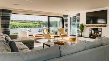 A modern living room with a large sofa, two armchairs, a coffee table, and a fireplace. A large window offers a view of greenery outside.