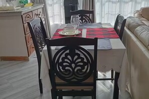 Un comedor con una mesa dispuesta para dos personas, con mantel rojo y negro, una copa y un plato. A la derecha hay un sofá de cuero y, al fondo, una zona de cocina.