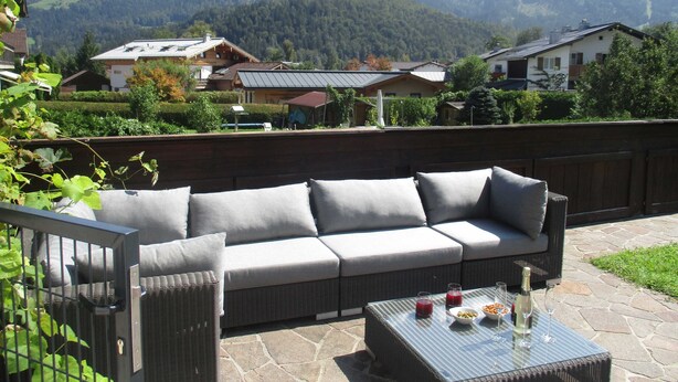 Un canapé en osier avec des coussins, une table en verre avec des boissons, et une vue sur les montagnes et les maisons.