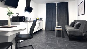 A modern living room with a grey sofa, a dining table with chairs, a kitchen area with a microwave, and a flat-screen TV mounted on the wall. - Apartment 'In Fuldatal - Kassel' with Private Garden and Wi-Fi (Fuldatal)